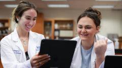 Two female students in lab coats smile as they stare at an iPad.