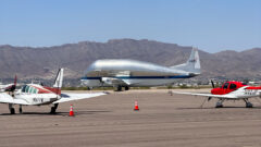 A massive silver Super Guppy aircraft taxies in a runway with mountains in the backdrop.