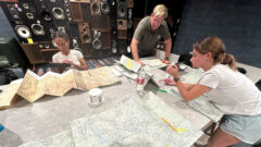 Marisa Vietti, Nathan Rogers and Jensen Kervern gather around a table filled with flight charts and maps.
