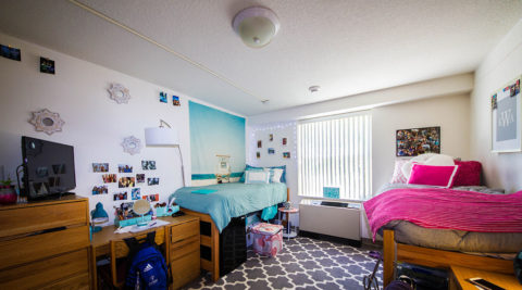 Two twin beds inside a Trinity dorm room, one with blue and one with pink. Student photos on the wall and a TV on a dresser.