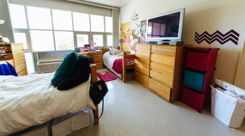 Dorm room with one dresser and TV, two twin-sized beds, and a large window in Lynn's Residence Center.