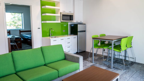 Perper Residence Hall living area including a green couch, coffee table, sink with green backsplash, fridge and dining table with green chairs.