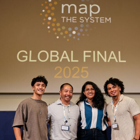 Associate Professor, Wayne Law, and Lynn students Ian Waite, Vishwa Shukla, and Daniel Miranda Duarte attend 2025 global final for Map of Systems competition. They take a group photo in front of a projector "Map the System: Global Final 2025."