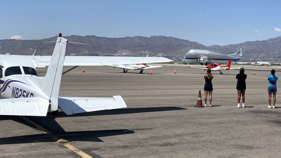 Flight instructors Blaise DaCosta, Jensen Kervern, and Marisa Vietti take a picture of a large silver airplane with mountains in the backdrop.