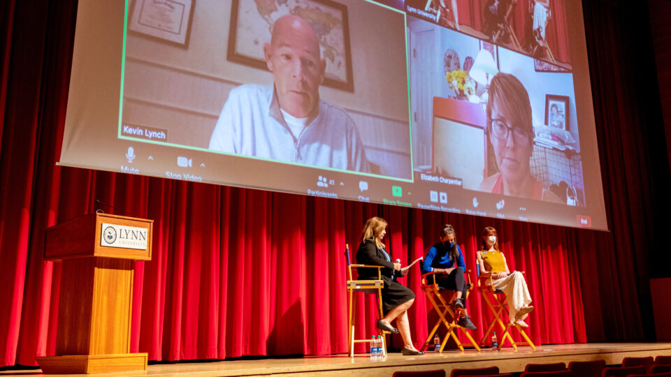 A panel of three and virtual guests appearing via Zoom are on stage in the Wold Performing Arts Center