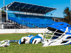 Lacrosse helmet, gloves, and stick in front of bleachers on the field.