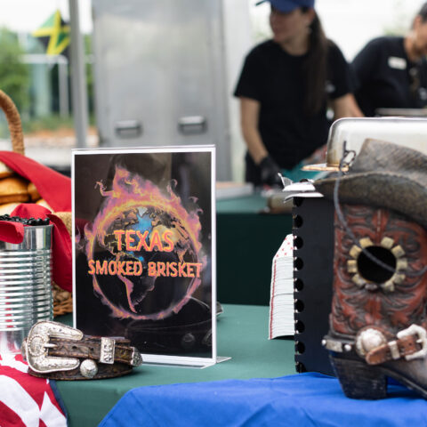 A table with signage that reads: "Texas Smoked Brisket". On the table, a U.S. Flag, a cowboy boot and a basket with bread buns.