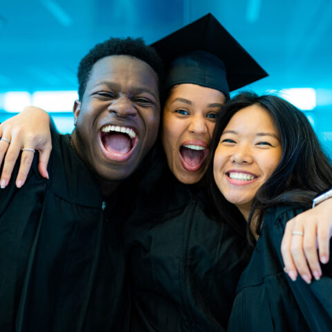 Three graduates, one with a graduation cap, pose for a photo together excited.