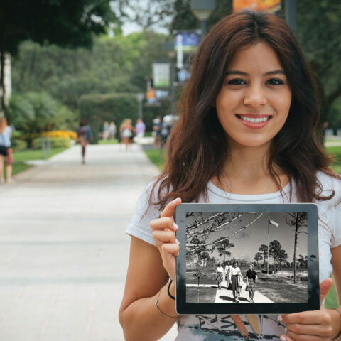 Student holds an iPad displaying a historical picture of campus while standing in the main walkway on campus.