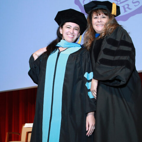 Elena McDonald takes a picture with Kathleen Weigel on stage in the Wold Performing Arts Center. Both wearing cap and gowns.