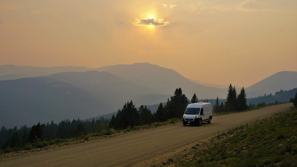A campervan drives down the road at sunset with a view of mountains in the background.
