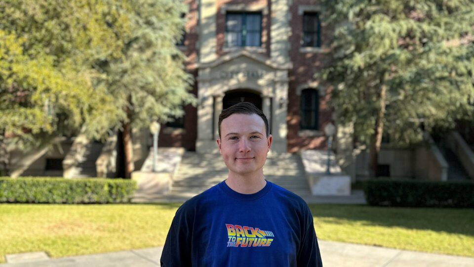 Chase stands outside of the set where the “Back to the Future” film was made and is wearing a shirt with the “Back to the Future” logo.